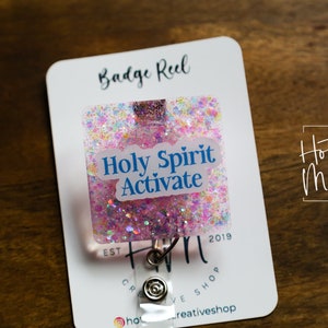 Może przedstawiać: Rolka do odznak z różowymi i białymi błyskotkami z białą naklejką z napisem "Holy Spirit Activate". Rolka jest przymocowana do białej karty z tekstem "Badge Reel" i "Hot Mess Creative Shop".