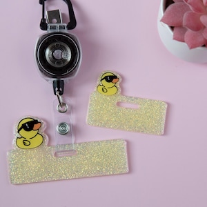 Puede incluir: Un portatarjetas retráctil con dos portaetiquetas brillantes. Cada soporte tiene un pato amarillo con gafas de sol. El portatarjetas es negro y plateado. El fondo es rosa claro.