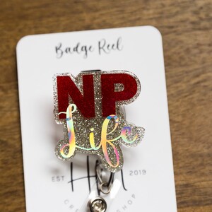 May include: A silver and red glitter badge reel with the text "NP life" on it. The badge reel is attached to a white card with the text "Badge Reel" and "Est. 2018" on it.