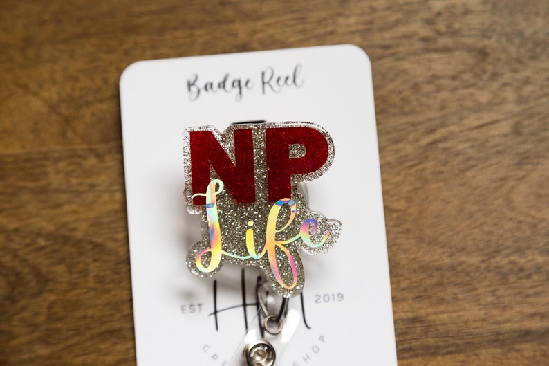 May include: A silver and red glitter badge reel with the text "NP life" on it. The badge reel is attached to a white card with the text "Badge Reel" and "Est. 2018" on it.