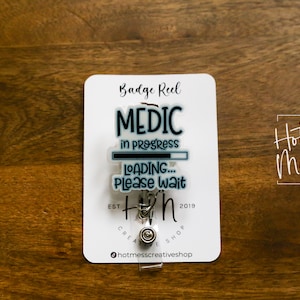 以下が含まれることがあります： 「MEDIC in progress LOADING... Please wait」と「Hot Mess Creative Shop」の文字が書かれた、青と黒のグラフィックが施された白いバッジリールです。