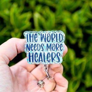 May include: A badge reel with the text "THE WORLD NEEDS MORE HEALERS" in blue lettering. The badge has a glittery, light blue border and a clear plastic reel with a metal clip. The background is blurred green foliage.
