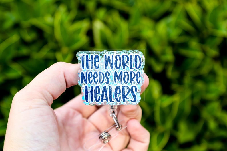 May include: A badge reel with the text "THE WORLD NEEDS MORE HEALERS" in blue lettering. The badge has a glittery, light blue border and a clear plastic reel with a metal clip. The background is blurred green foliage.