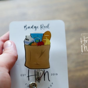 May include: A brown paper bag with a white handle filled with groceries, including a carton of milk, a blue juice box, a loaf of bread, a banana, an orange, and an apple. The bag is attached to a retractable badge reel with a gold metal clip. The badge reel is on a white card with the text "Badge Reel" and "Hot Mess Creative Shop" printed on it.