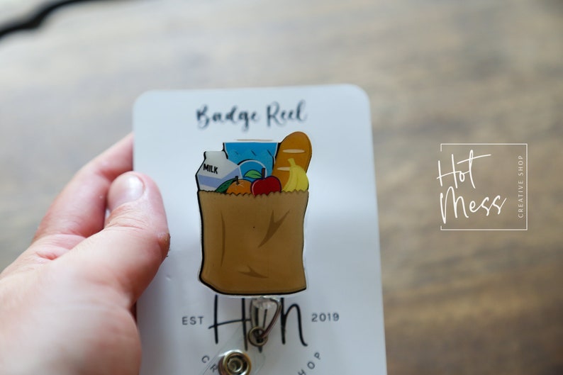 May include: A brown paper bag with a white handle filled with groceries, including a carton of milk, a blue juice box, a loaf of bread, a banana, an orange, and an apple. The bag is attached to a retractable badge reel with a gold metal clip. The badge reel is on a white card with the text "Badge Reel" and "Hot Mess Creative Shop" printed on it.