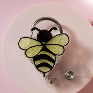 May include: A bee-shaped retractable badge holder with a glittery yellow and black design. The badge reel has a silver metal clip and a clear plastic strap for attaching to clothing. The bee's wings sparkle, and the body has black stripes.