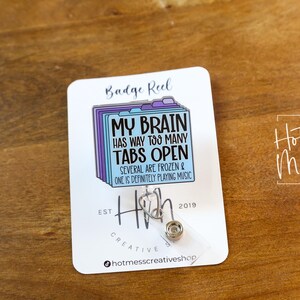 May include: A white badge reel with the text "MY BRAIN HAS WAY TOO MANY TABS OPEN" in black and blue. The reel features a design of file folders and a retractable cord. The reel is on a wooden surface.