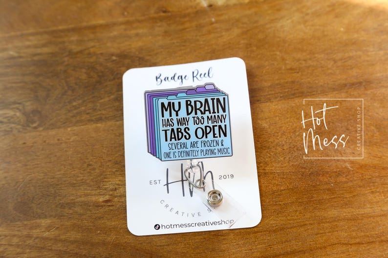 May include: A white badge reel with the text "MY BRAIN HAS WAY TOO MANY TABS OPEN" in black and blue. The reel features a design of file folders and a retractable cord. The reel is on a wooden surface.