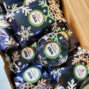 May include: A collection of small rectangular packages wrapped in dark blue paper with a white snowflake pattern. Each package features a circular sticker with the text Open December followed by the specific date, or Merry Christmas with a tree.