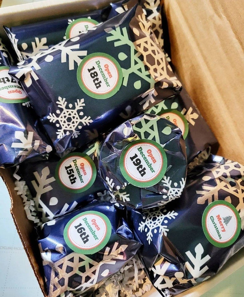 May include: A collection of small rectangular packages wrapped in dark blue paper with a white snowflake pattern. Each package features a circular sticker with the text Open December followed by the specific date, or Merry Christmas with a tree.