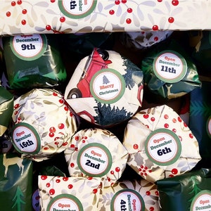 May include: A collection of small gifts wrapped in festive paper featuring red berries, green leaves, and a red truck illustration. Each package has a circular sticker with text such as Merry Christmas or Open December followed by a specific date.