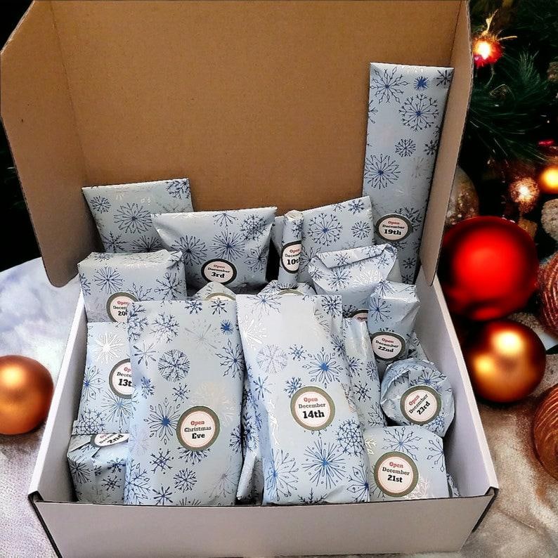 May include: An open cardboard box filled with various small packages wrapped in light blue paper featuring a dark blue snowflake pattern. Each package has a circular sticker with text indicating specific dates in December to open.