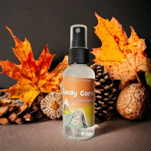 May include: A clear glass bottle with a black spray top and a white label that says "Forevershine Collections Candy Corn Room Spray". The label has a cartoon image of a candy corn and a pile of corn kernels. The bottle is sitting on a table with fall leaves and pine cones.