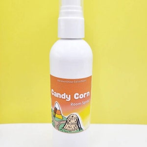 May include: White bottle of Candy Corn Room Spray with a cartoon image of a candy corn pile and a ladder. The label is orange and yellow with the text "Forevershine Collections" and "Candy Corn Room Spray".