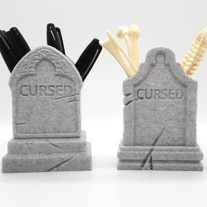 May include: Two gray tombstone-shaped desk organizers. One has black pens inside and the other has white plastic bones. Both tombstones have the word "CURSED" engraved on them.