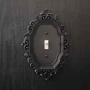 May include: A black light switch with an ornate, decorative frame. The frame is made of metal and has a scroll-like design.
