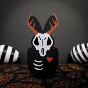 May include: Black wooden rabbit figurine with a white skull, red heart, and skeletal ribs. The rabbit has large, brown antlers.