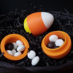 May include: A Halloween still life featuring a large candy corn and two small orange containers filled with white and brown sweets. The items are set against a backdrop of black shredded paper, creating a festive and colourful display.