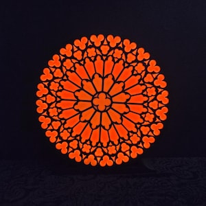 Può includere: Un design di vetrata arancione illuminata su sfondo scuro. Il motivo circolare presenta un motivo floreale centrale circondato da elementi a forma di petalo ripetuti, creando un design simmetrico e ornato.