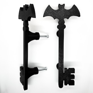 May include: Two black metal key-shaped door handles. One handle features a bat silhouette at the top, while the other has a stylized shape. Each handle has a screw for mounting. The handles are against a white background.