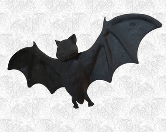 Może przedstawiać: Czarna figurka nietoperza z rozłożonymi skrzydłami. Nietoperz wykonany jest z gładkiego, ciemnego materiału i ma szczegółową twarz.