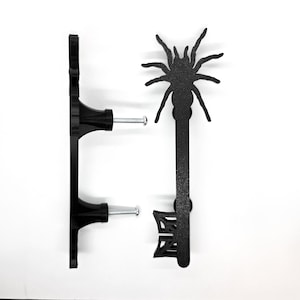 May include: Black metal cabinet pulls. One pull is a standard handle, the other is a key-shaped pull with a spider design at the top. Both pulls have screws for installation. The pulls are set against a white background.