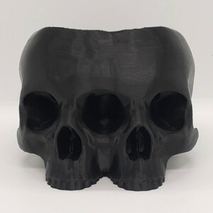 May include: A black 3D printed skull planter with three eye sockets and a detailed design. The planter is made of plastic and has a smooth, matte finish.