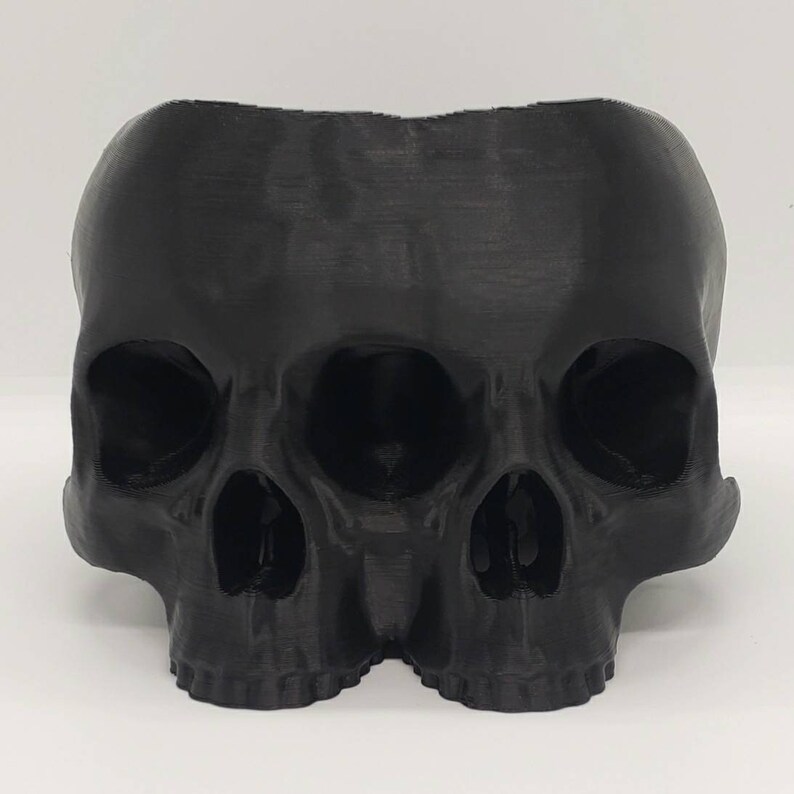 May include: A black 3D printed skull planter with three eye sockets and a detailed design. The planter is made of plastic and has a smooth, matte finish.