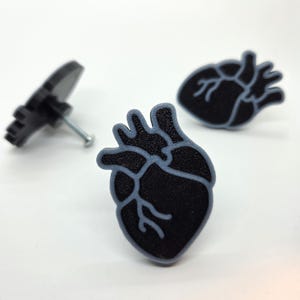 May include: Three black heart-shaped cabinet knobs with grey outlines and a silver screw. The knobs are arranged on a white surface. The knobs are a decorative element for furniture.