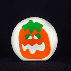 Puede incluir: Una luz redonda blanca con una cara de calabaza naranja de dibujos animados con un tallo verde y ojos y dientes blancos. La calabaza está iluminada desde atrás, haciendo que la luz brille.