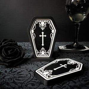Könnte beinhalten: Schwarze und weiße Untersetzer in Sargform mit Kreuzdesign. Die Untersetzer sind aus Holz und haben eine glänzende Oberfläche.