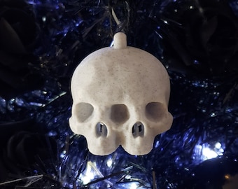 Puede incluir: Adorno de calavera de cerámica blanca con superficie texturizada. El adorno tiene forma de cráneo humano con dos cuencas de los ojos, una nariz y una boca.