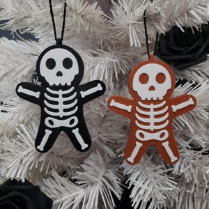 Könnte beinhalten: Zwei Lebkuchen-Anhänger in Skelettform, einer schwarz und einer orange, hängen an einem weißen künstlichen Weihnachtsbaum.