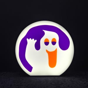 Puede incluir: Un fantasma blanco con un halo morado y una sonrisa naranja, en forma de círculo. El fantasma está saludando con su mano izquierda.