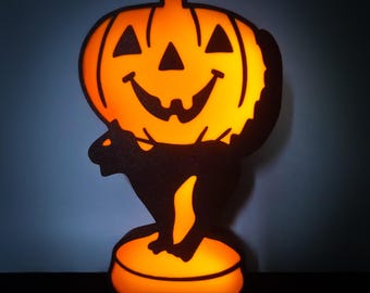 May include: A black cat silhouette holding a glowing orange jack-o'-lantern with a smiling face. The cat is standing on a round orange base.