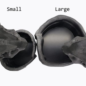 Puede incluir: Dos macetas de cer&aacute;mica negra con forma de calavera y parte superior abierta. La maceta de la izquierda est&aacute; etiquetada como "Peque&ntilde;a" y la maceta de la derecha est&aacute; etiquetada como "Grande".