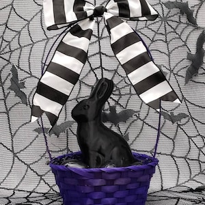 May include: A black rabbit figurine sits in a purple wicker basket with a black and white striped bow on top. The basket is on a black and white spiderweb background.