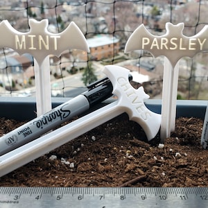 May include: Three white plastic bat-shaped plant markers with the words "MINT", "PARSLEY", and "CHIVES" written on them. Two black Sharpie permanent markers are lying on the ground next to the markers.