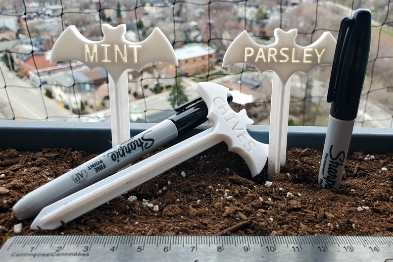 May include: Three white plastic bat-shaped plant markers with the words "MINT", "PARSLEY", and "CHIVES" written on them. Two black Sharpie permanent markers are lying on the ground next to the markers.