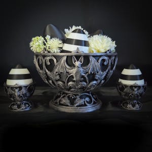 Puede incluir: Un centro de mesa decorativo con un cuenco ornamental negro y plateado con un diseño de murciélago, que contiene objetos en forma de huevo a rayas blancas y negras y flores blancas. Dos portahuevos más pequeños a juego flanquean el cuenco.