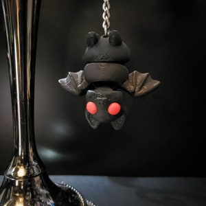 May include: A black bat-shaped charm with red eyes hangs from a silver chain. The charm is made of polymer clay and has a textured surface.