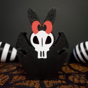May include: Black and white skull with red bunny ears and wings. The skull has a black outline and white eyes. The wings are black and have a textured surface. The bunny ears are black and have a red bow on top.
