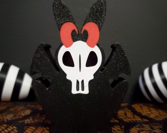 Pode incluir: Crânio preto e branco com orelhas de coelho vermelhas e asas. O crânio tem um contorno preto e olhos brancos. As asas são pretas e têm uma superfície texturizada. As orelhas de coelho são pretas e têm um laço vermelho no topo.