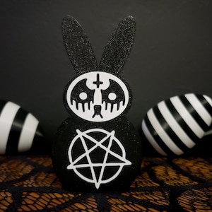 May include: Black and white bunny rabbit figurine with a pentagram and a skull with a cross on its head. The bunny is sitting on a black and white patterned surface.
