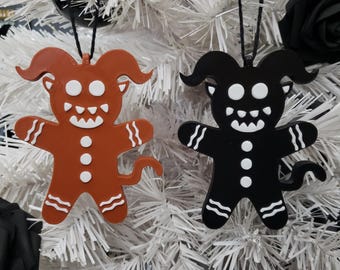 Puede incluir: Dos adornos navideños de estilo gingerbread, uno rojo y otro negro, con detalles blancos. Ambos adornos presentan una cara diabólica con cuernos y cola.