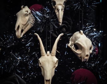 Puede incluir: Una colección de adornos de calaveras blanquecinas, que incluyen una calavera de caballo, de pájaro y de cabra, cuelgan de un árbol de Navidad de oropel negro. Adornos rojos se intercalan entre las calaveras. Los adornos están suspendidos por cuerdas negras.