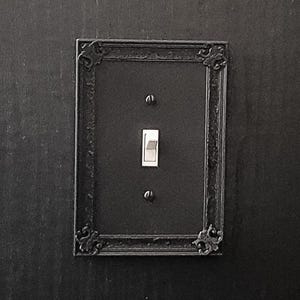 Könnte beinhalten: Eine schwarze Lichtschalterplatte mit einem verzierten Rahmen und einem einzigen Kippschalter.