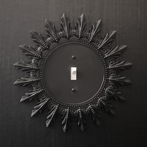 May include: A black light switch plate with a decorative sunburst design. The switch plate is made of metal and has a smooth, matte finish.
