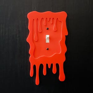 Könnte beinhalten: Ein roter Lichtschalter-Abdeckung in Form einer tropfenden Flüssigkeit. Die Abdeckung ist so konzipiert, dass sie wie Blut aussieht, das an einer Wand heruntertropft.