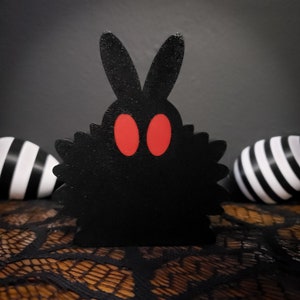 May include: Black bunny rabbit figurine with red eyes and a fuzzy texture. The bunny is sitting on a black lace surface.